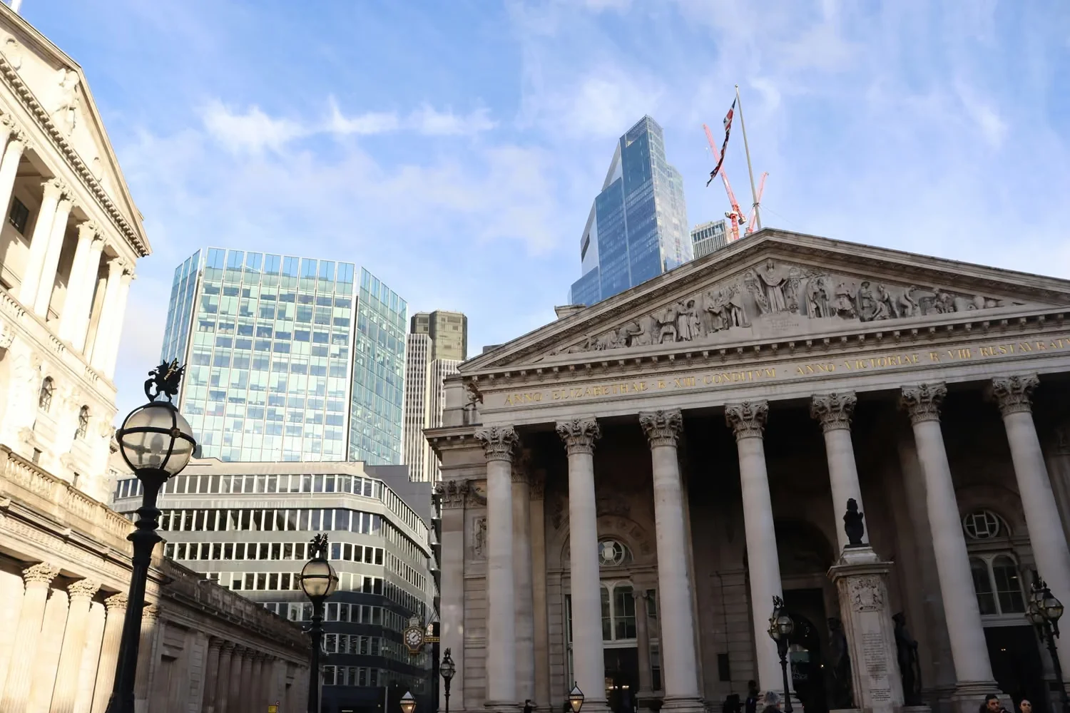 A low-angle photograph capturing London financial architecture, featuring a classical stone building with columns in the foreground and modern glass skyscrapers behind it.