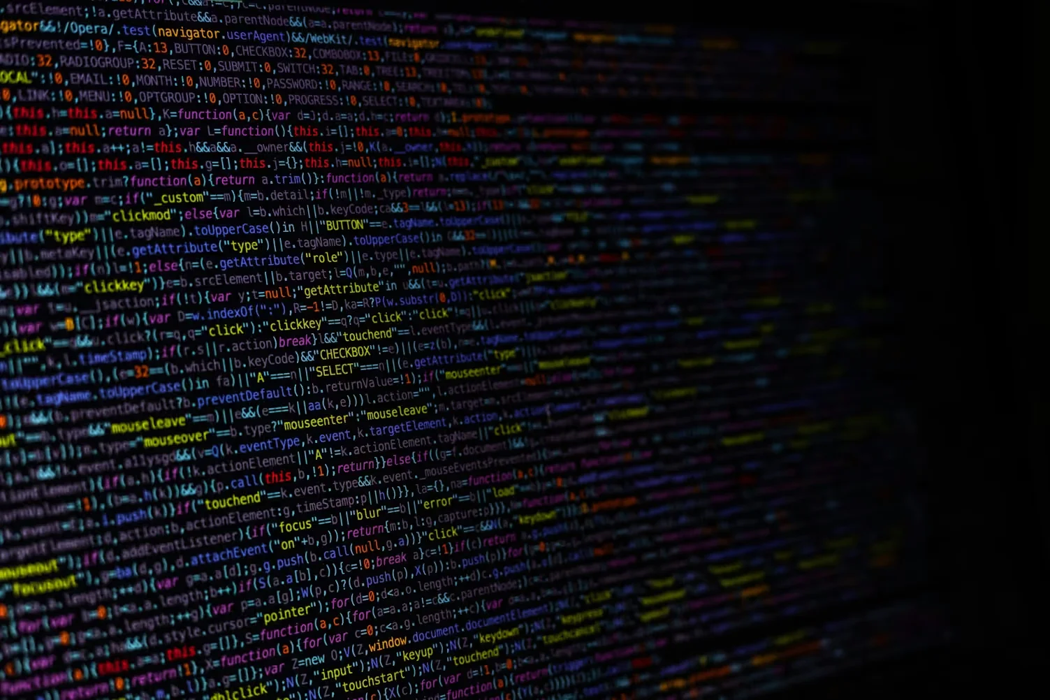 A close-up photograph of lines of colourful programming code displayed on a dark computer screen, with a shallow depth of field blurring the edges.