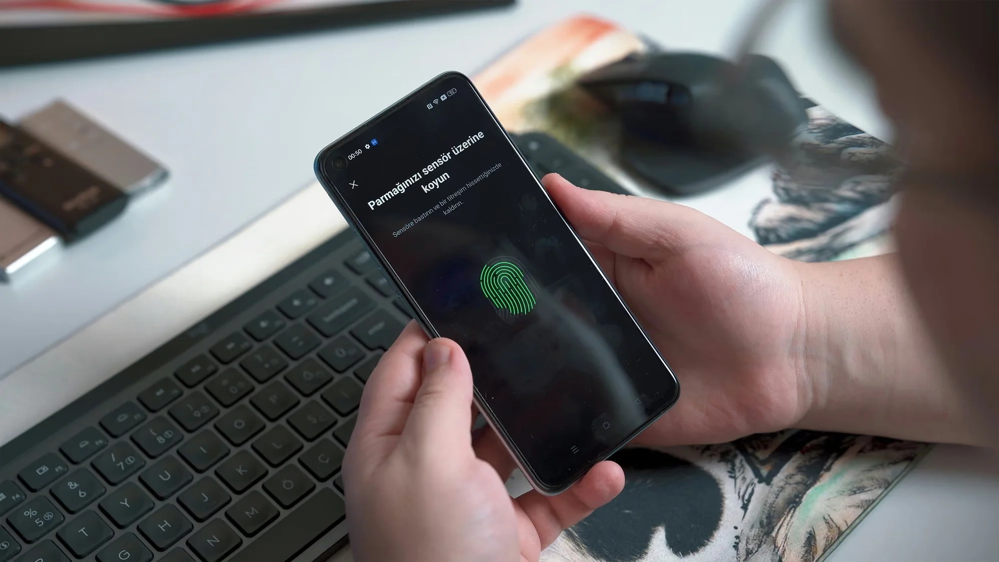 A person is holding a smartphone displaying a fingerprint scanner setup prompt with Turkish text on the screen, sitting at a desk with a keyboard and mouse.